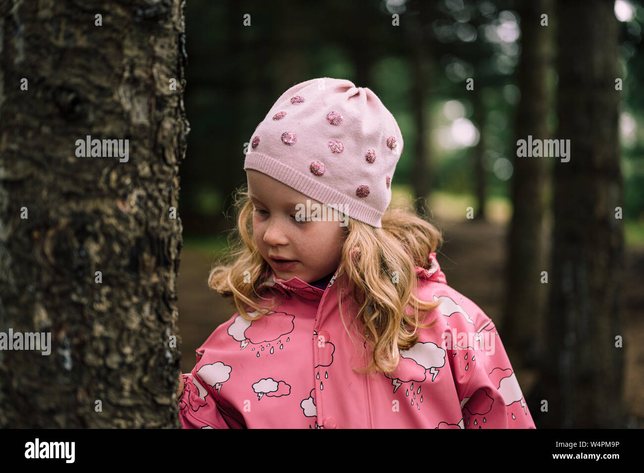 Blonde haired girl in pink raincoat and hat standing next to tree in Reykjavik, Iceland forest Stock Photo