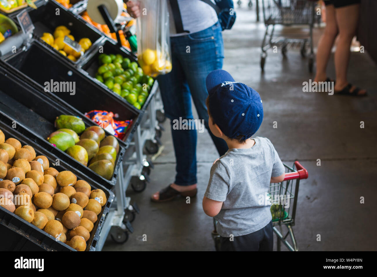 Curious toddler pusing small cart shopping with mother at farmers market. Stock Photo