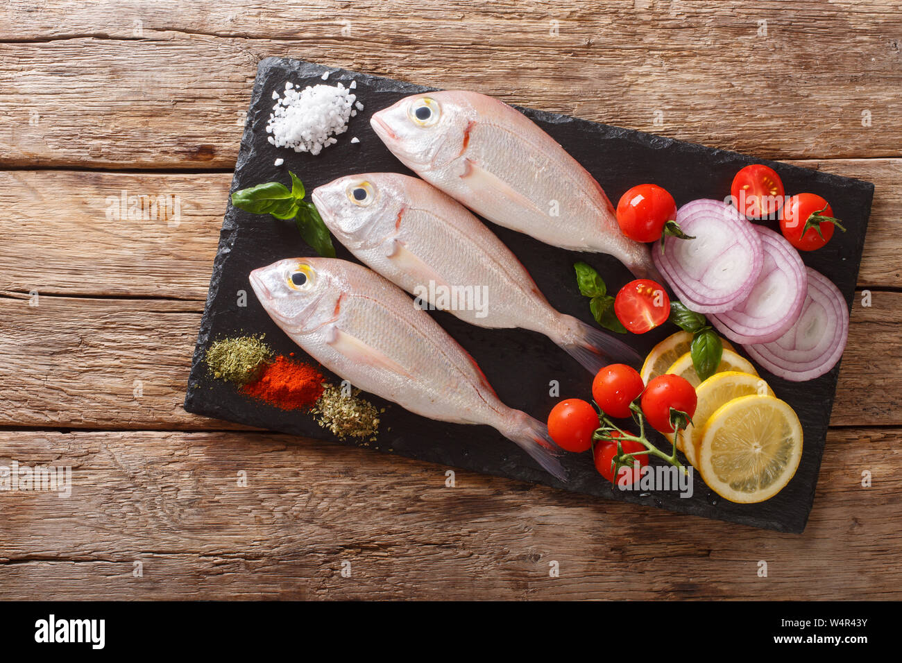 Uncooked dorado or gilt-head sea bream fish with spices, tomato, onion and lemon close up on a slate board on the table. horizontal top view from abov Stock Photo