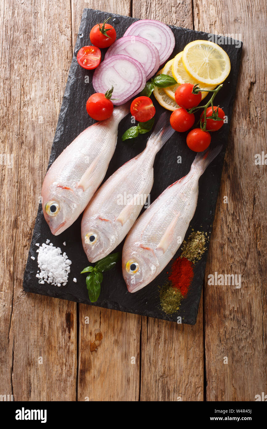 Uncooked dorado or gilt-head sea bream fish with spices, tomato, onion and lemon close up on a slate board on the table. Vertical top view from above Stock Photo