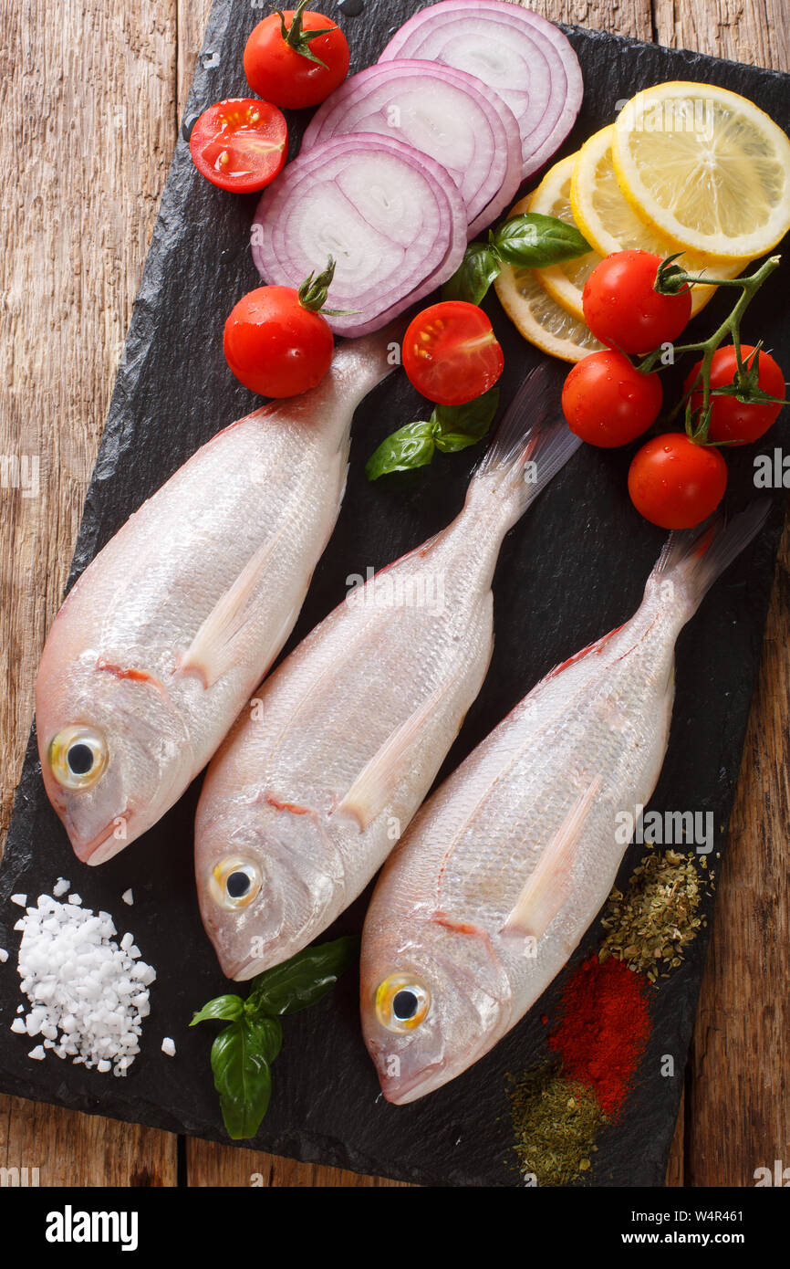 Raw pink dorado or gilt-head sea bream fish with spices, vegetables and lemon close-up on a slate board on a table. Vertical top view from above Stock Photo