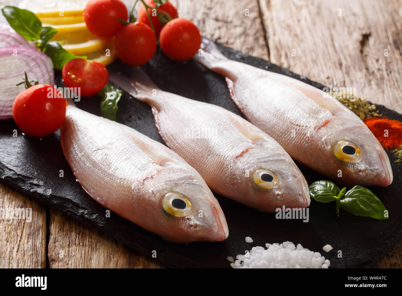 Uncooked dorado or gilt-head sea bream fish with spices, tomato, onion and lemon close up on a slate board on the table. horizontal Stock Photo