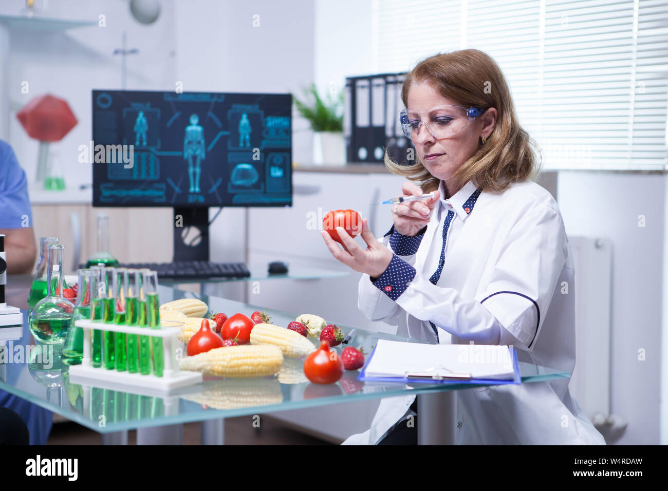 Biologist in white coat in a research lab injecting liquid in a tomato. Test tubes with green liquid. Stock Photo