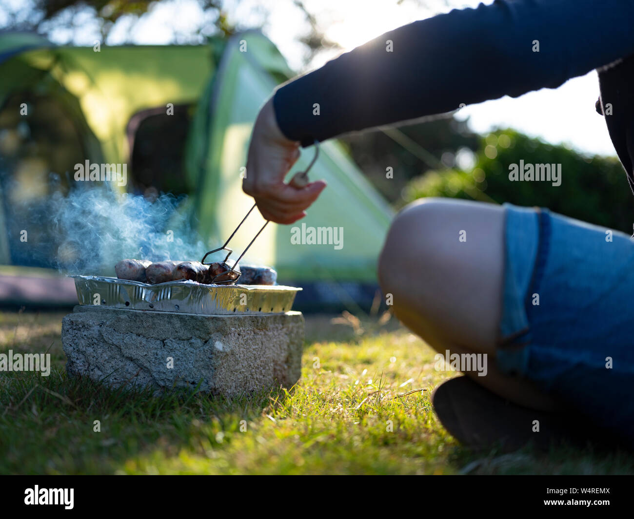 A young woman cooking sausages on a camping holiday, on Isles of Scilly, UK. Stock Photo