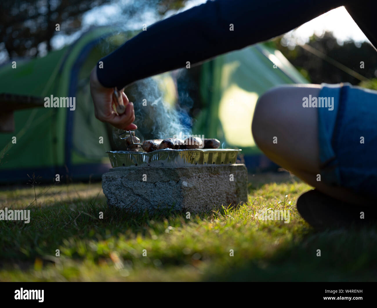 A young woman cooking sausages on a camping holiday, on Isles of Scilly, UK. Stock Photo