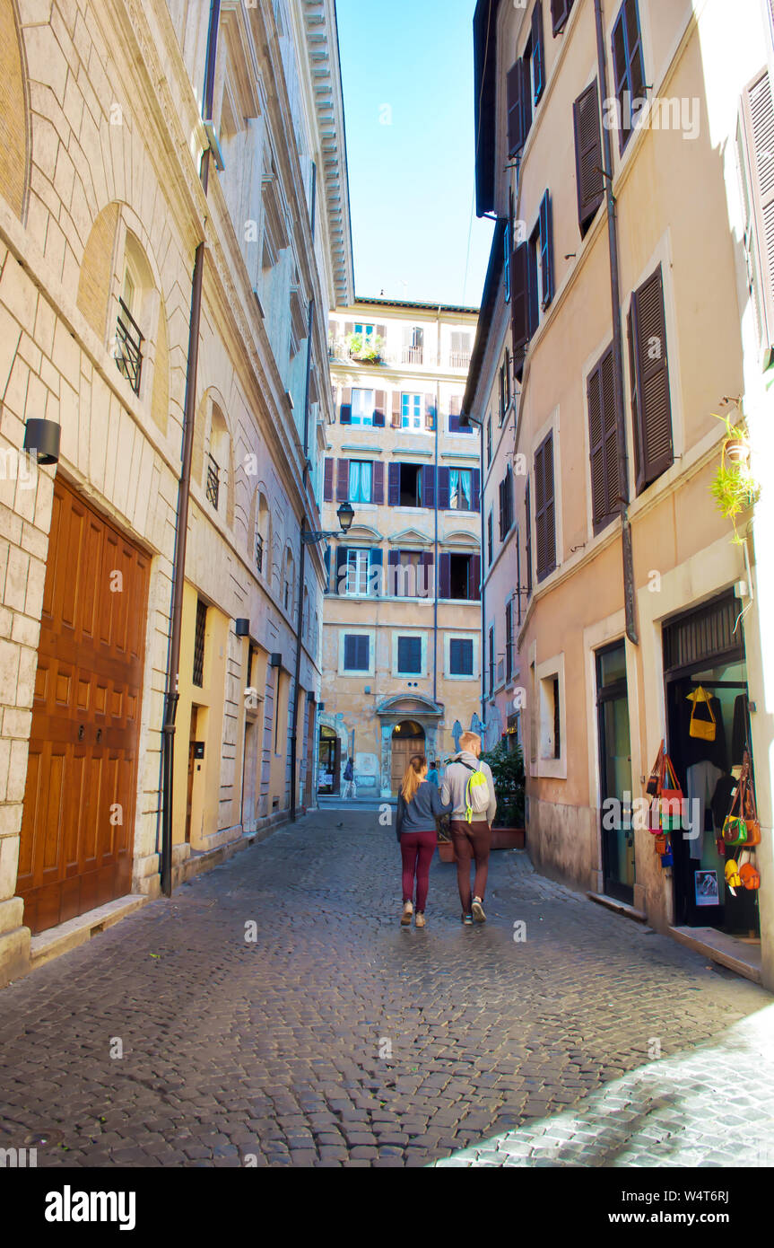 Young man and woman walking down a narrow solitary street in the city center of the capital of Italy, Rome. Warm fall morning, vibrant blue sky, calm Stock Photo