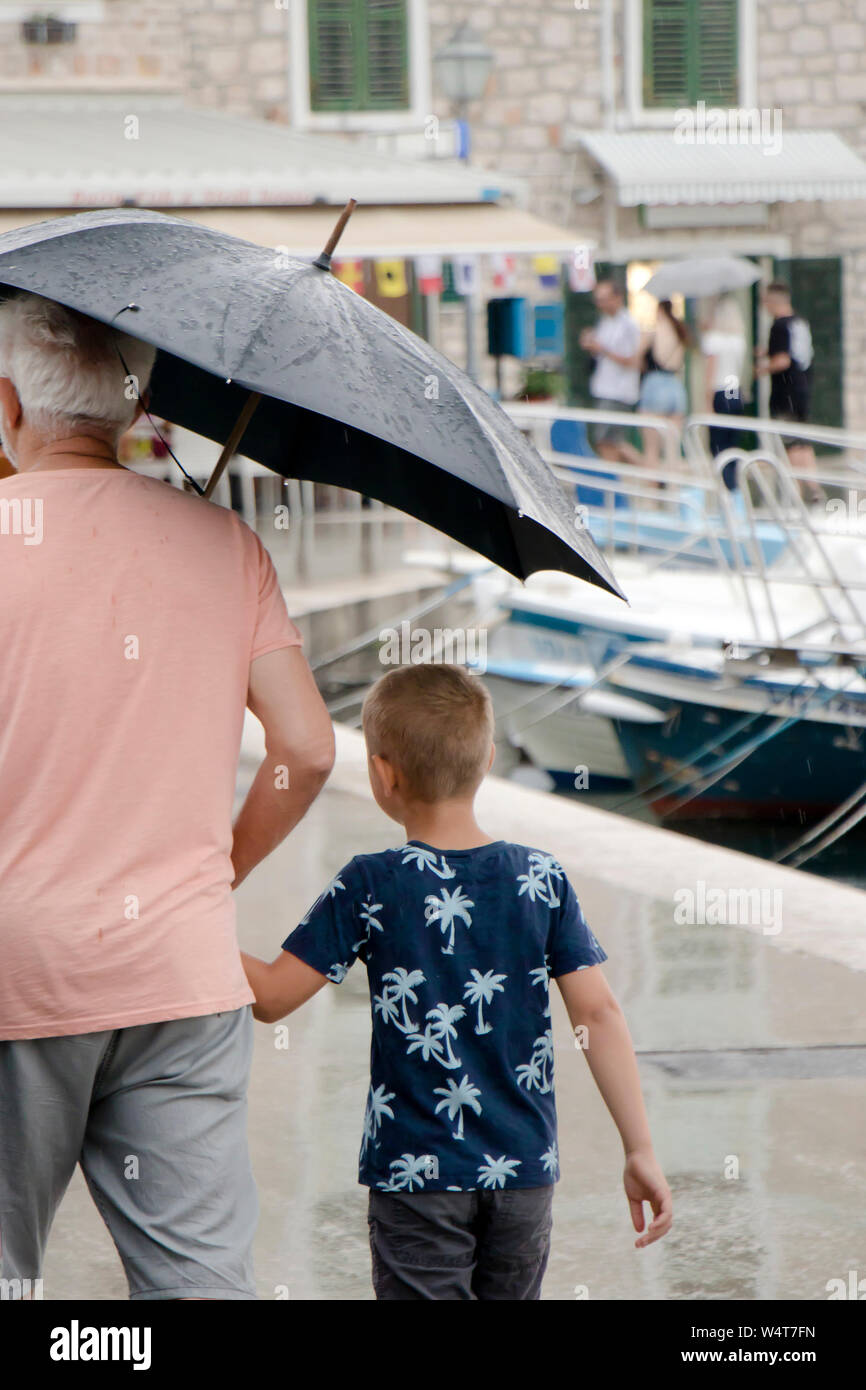 Vodice, Croatia - July 13, 2019: Boy and his grandfather walking under umbrella in on a rainy summer day by the sea pier Stock Photo