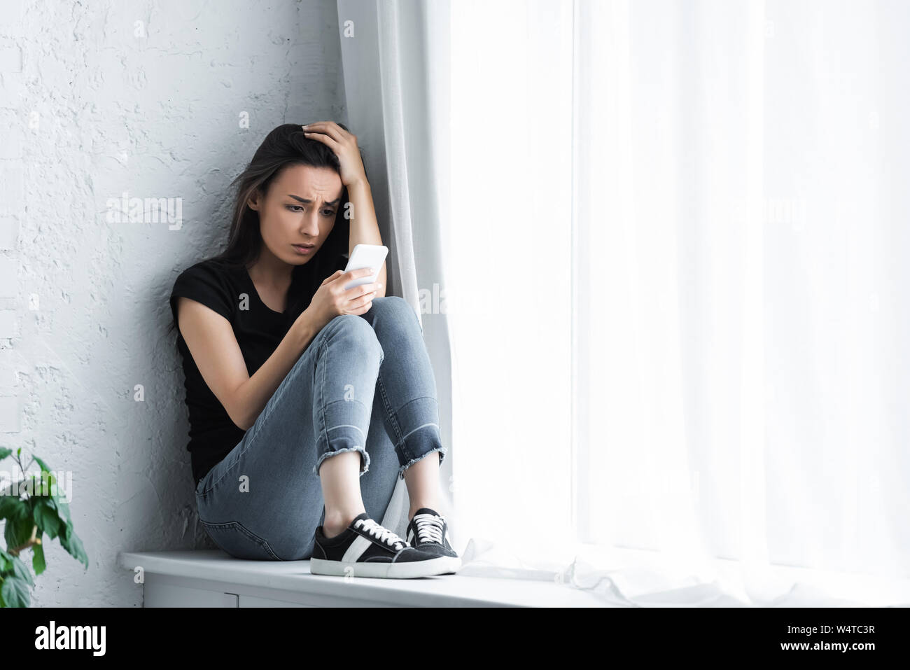 depressed young woman sitting on window sill at home and using smartphone Stock Photo
