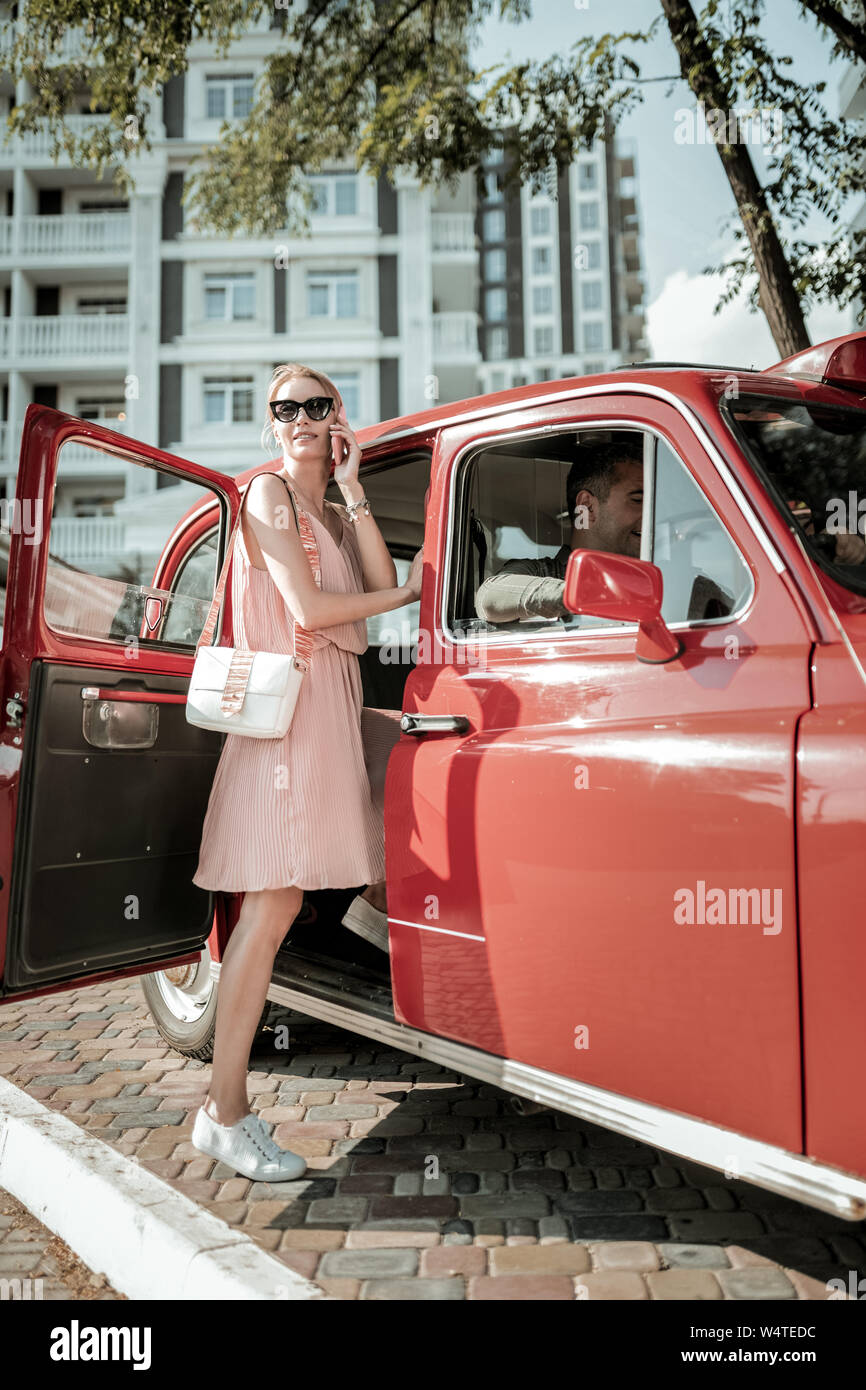 Great talker. Smiling pretty woman talking on her phone getting into the red retro-car before the car ride. Stock Photo