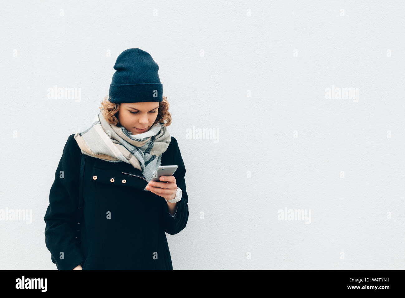 Young woman smiling and typing on mobile device over gray background with copy space. Hipster female using smart phone outdoors near wall in winter we Stock Photo