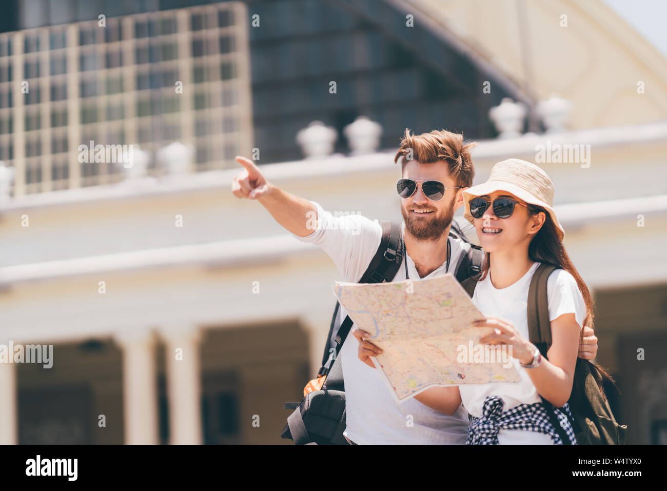Multi-ethnic traveler couple using local map together on sunny day, man pointing forward to copy space. Honeymoon trip, backpacker tourist, Asia city Stock Photo