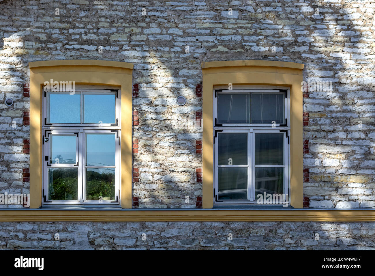 Two rectangular windows with yellow wooden frames on the background of a stone wall with a shadow from the tree. From the window series of the world. Stock Photo