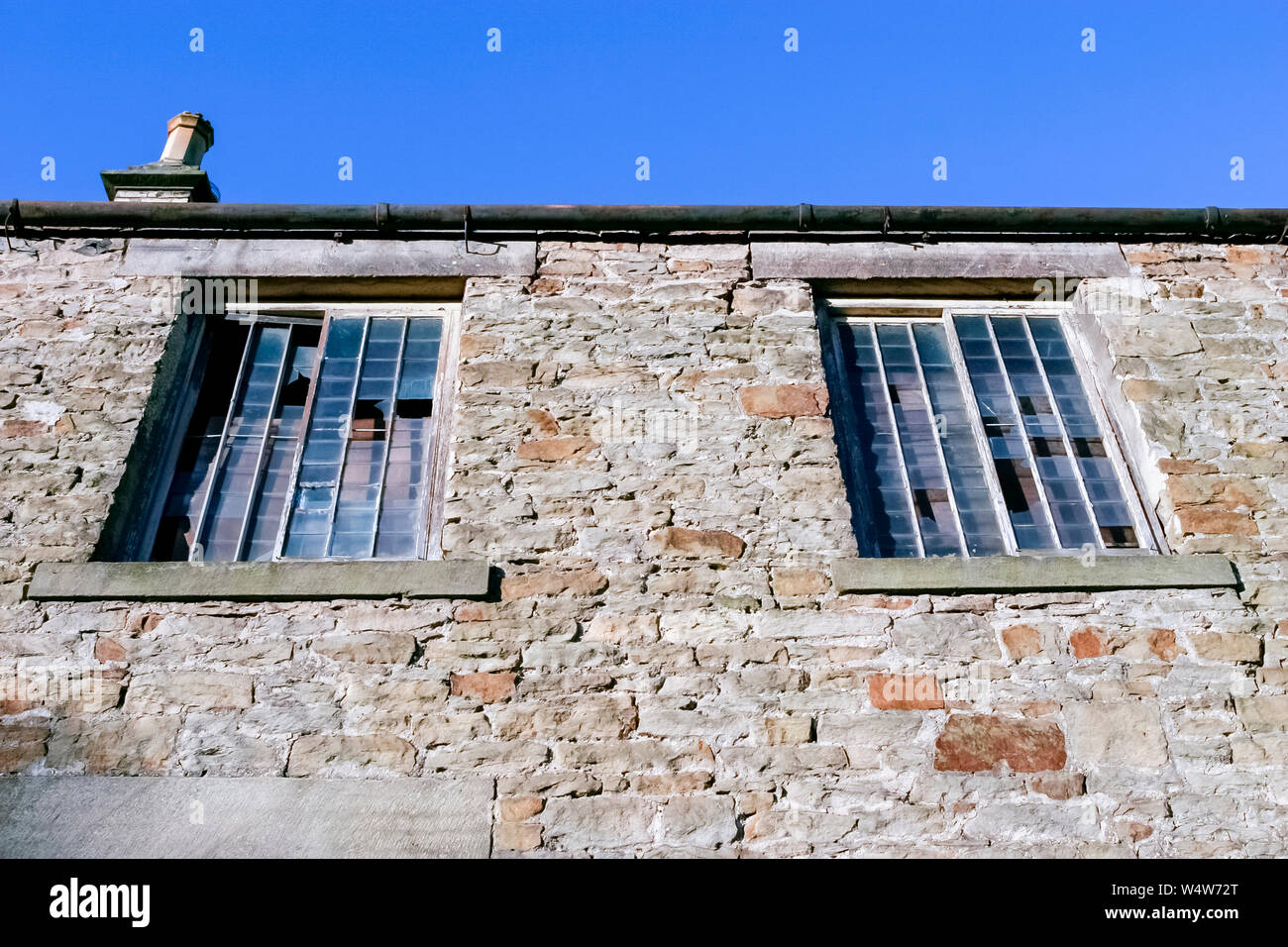 Looking up at an old derelict stone building with two barred and broken windows. Stock Photo