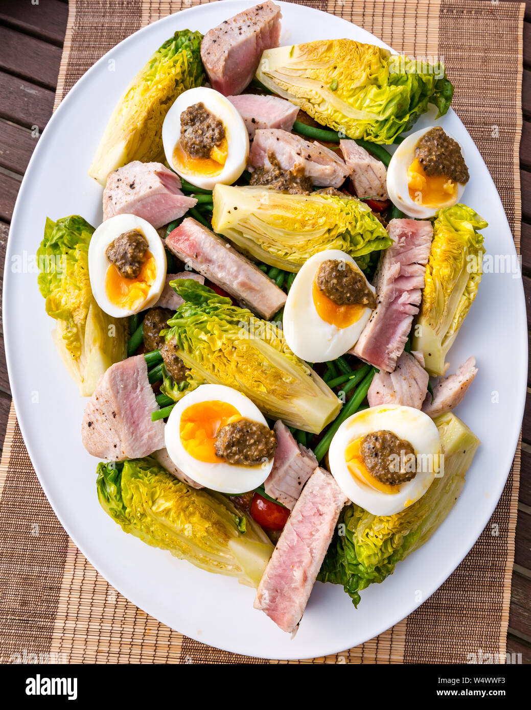 Version of salad Nicoise on white plate.  Hard boiled eggs and olive dressing, baby gem lettuce, boiled potatoes & tuna steak slices Stock Photo