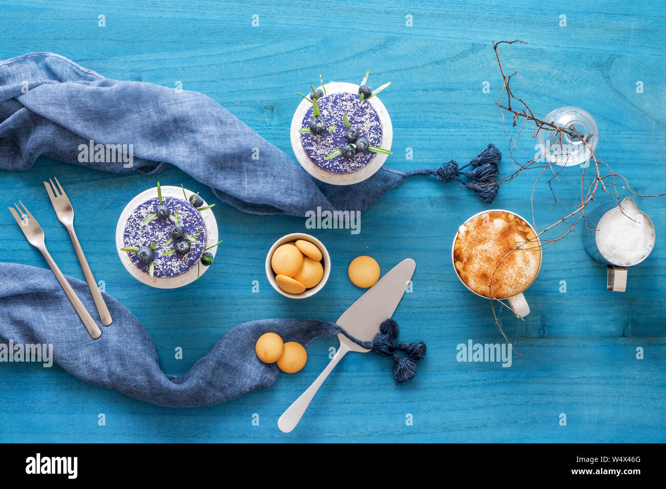 Two round no bake blueberry mini cheesecakes enveloped in coconut and embellished with real blueberries and rosemary leaves. Blue wooden table with co Stock Photo