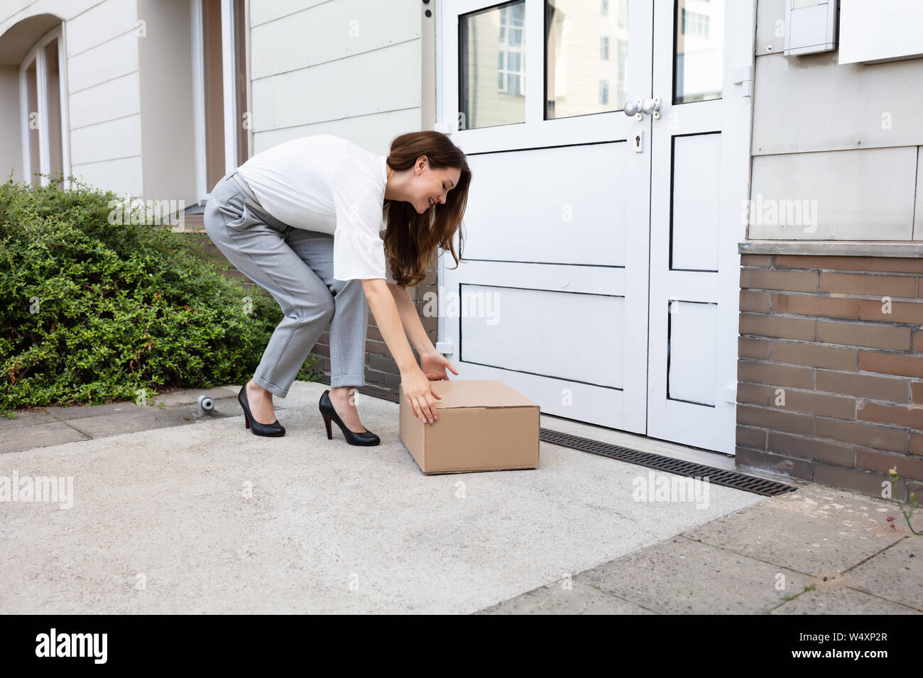 Smiling Young Woman Peeking Up The Delivered Parcel At The Door Entrance Stock Photo