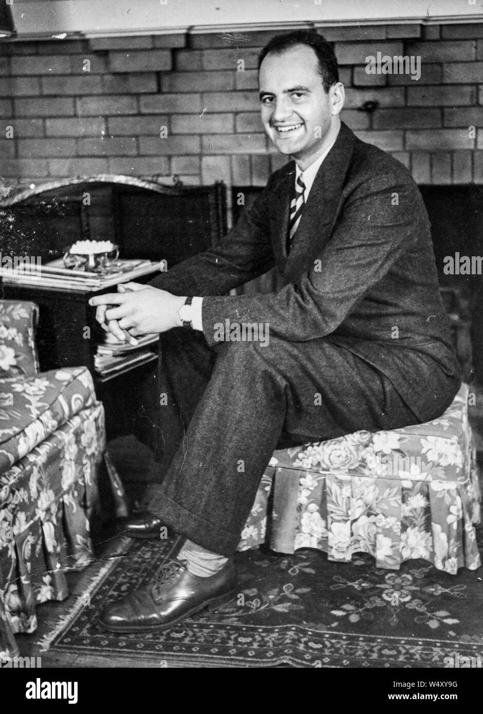 Full length portrait of a smiling man wearing a dark suit, leather shoes, a necktie, and a wristwatch, profile view with hands clasped, sitting on a floral print ottoman in front of a fireplace in a living room, Detroit, Michigan, 1960. () Stock Photo