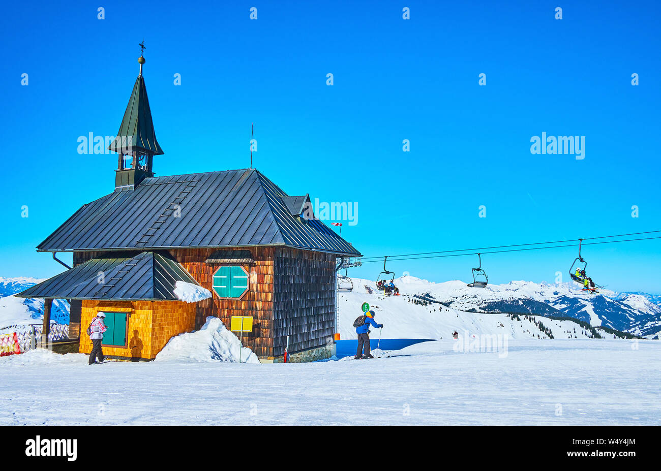 The snowy Alpine scenery and riding chairlift with sportsmen behind the old wooden Elisabeth chapel, located atop the Schmitten mount, Zell am See, Au Stock Photo