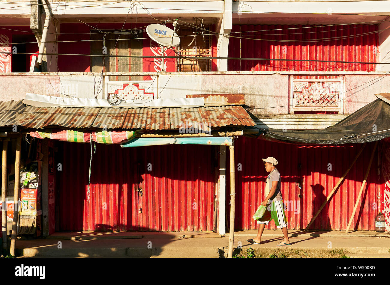 Cuartero, Capiz Province, Philippines: Young man walking in front of stores, closed with red metal folding doors, in late afternoon Stock Photo