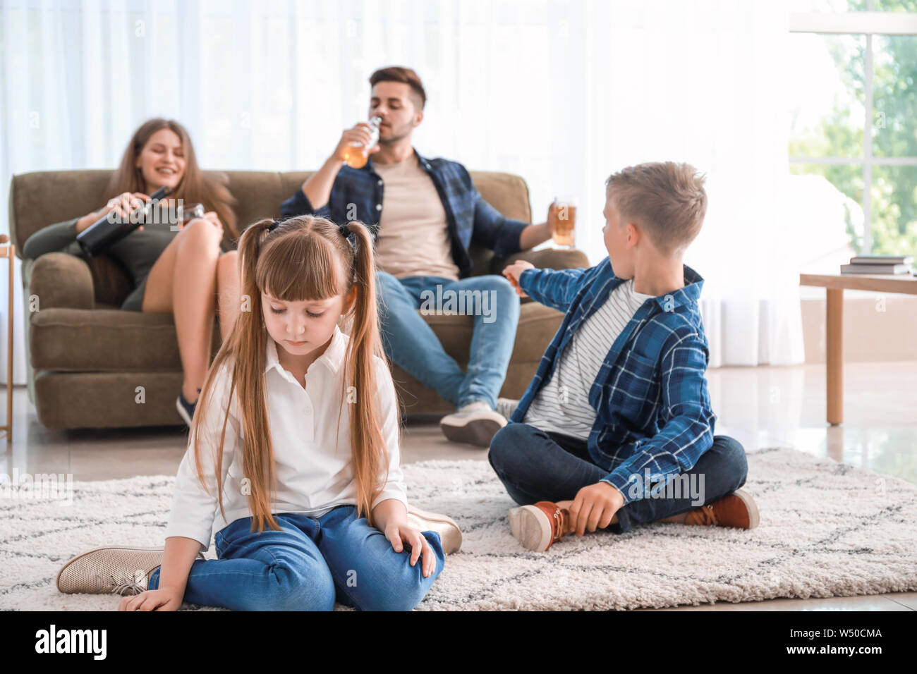 Sad little children sitting on floor while addicted parents drinking alcohol at home Stock Photo