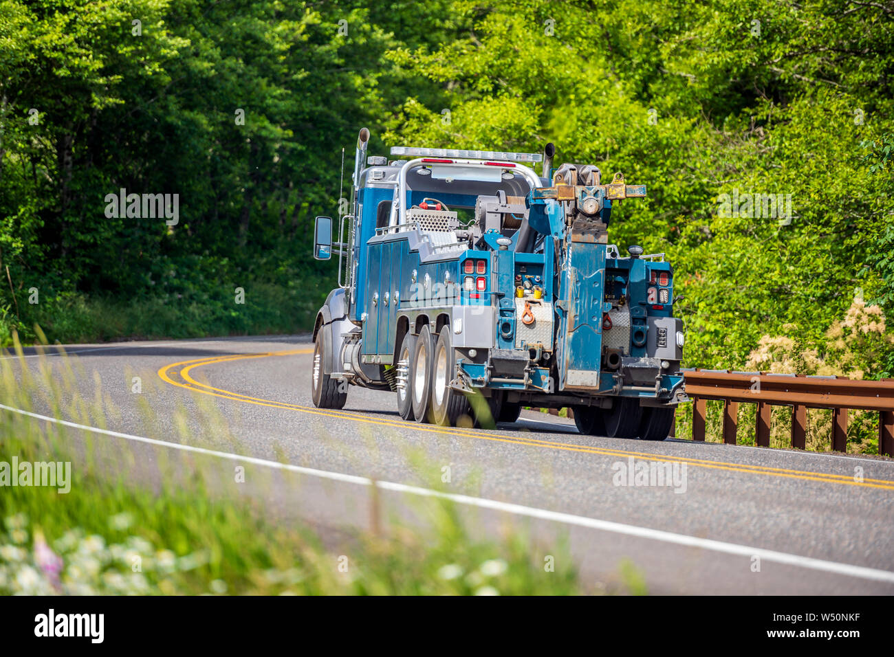 Powerful professional blue big rig towing semi truck for tow semi trucks running on the winding road in green forest for pick up a broken truck that n Stock Photo