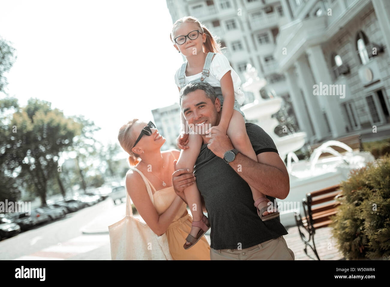 High spirits. Cheerful little girl riding her smiling fathers shoulders with her mother standing behind her. Stock Photo