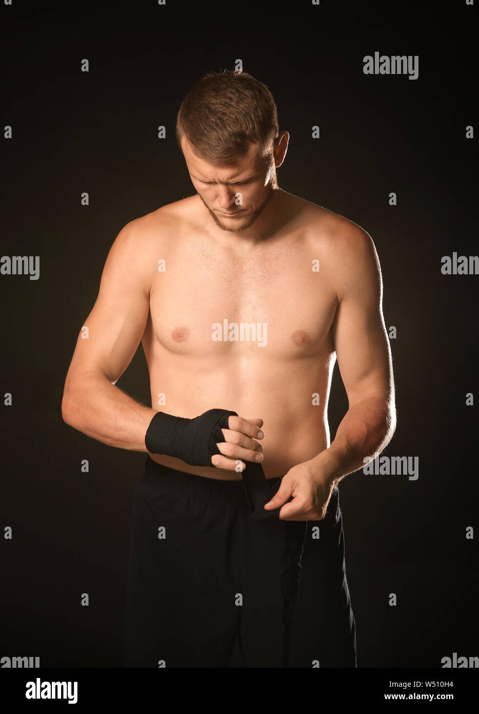 Strong male boxer applying wrist bands against dark background Stock
