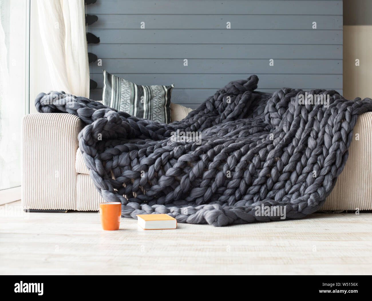 Merino wool grey blanket lying on white sofa with an orange cup and book near it Stock Photo