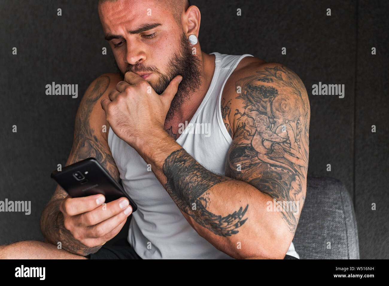 handsome man with tattoos and beard in white t shirt using smartphone and looking thoughtful at it W516NH