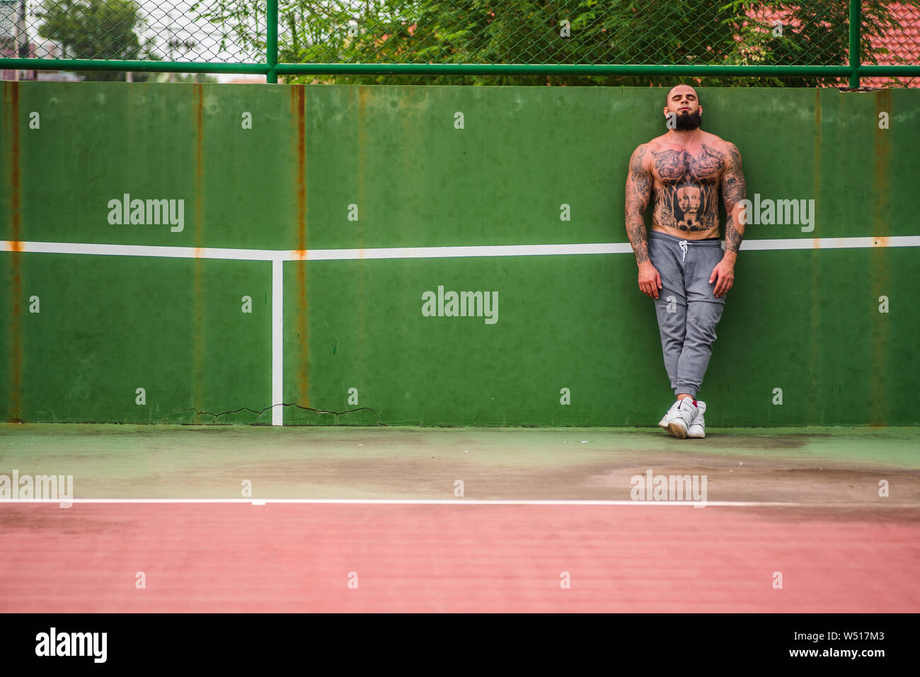 Attractive man with tattoos and beard wearing training pants standing on tennis court looking front Stock Photo