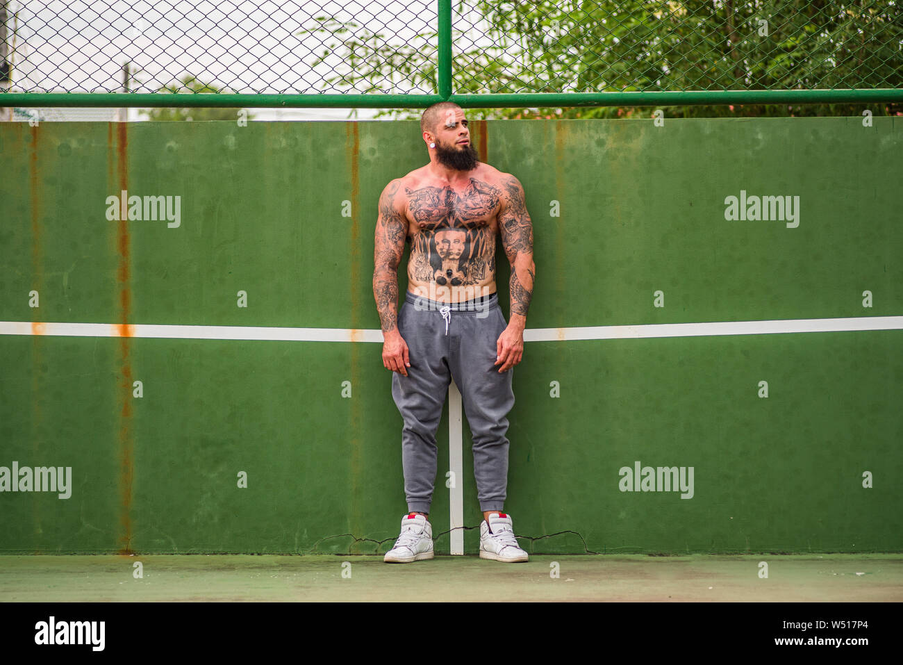 Young man with tattoos and beard wearing training pants and no shirt leaning on tennis court wall and looking to side Stock Photo
