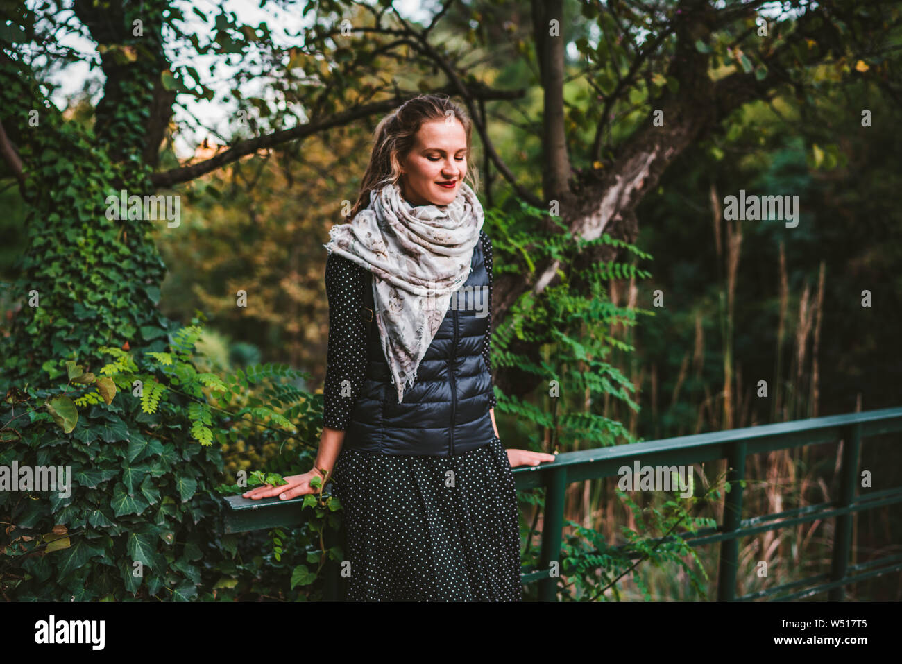 Pretty young woman standing on bridge in green park looking down and ...