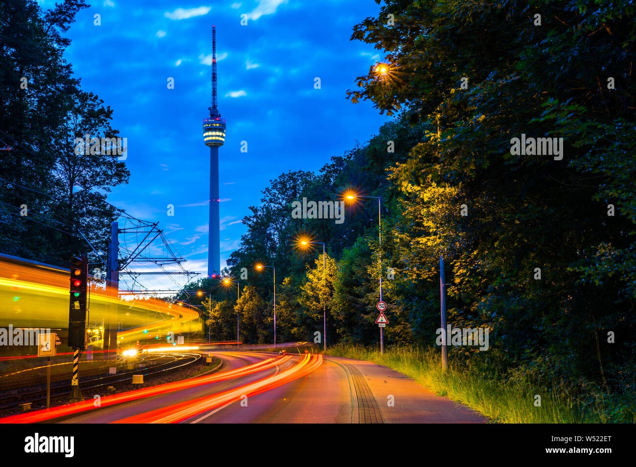Germany, Lights of illuminated train and traffic passing by street road to television tower of stuttgart city inside green forest nature landscape in Stock Photo