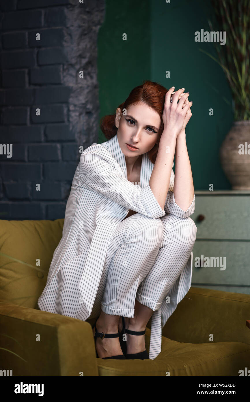 Concept: fashion, beauty. Pretty redhead thin lean young woman with vintage hairdo alluring in white unbuttoned pant suit. Studio photoshoot. Stock Photo