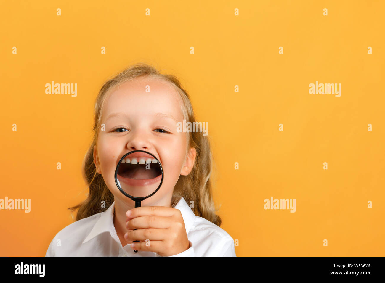 Portrait of a little girl schoolgirl on a yellow background. A child shows his teeth and smile through a magnifying glass. Stock Photo