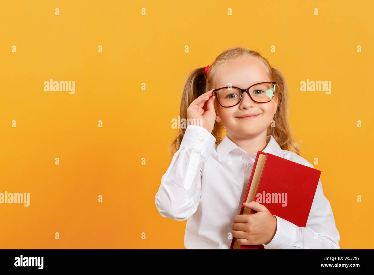 Portrait of a cute little girl on a yellow background. Child schoolgirl looking at the camera, holding a book and straightens glasses. The concept of Stock Photo