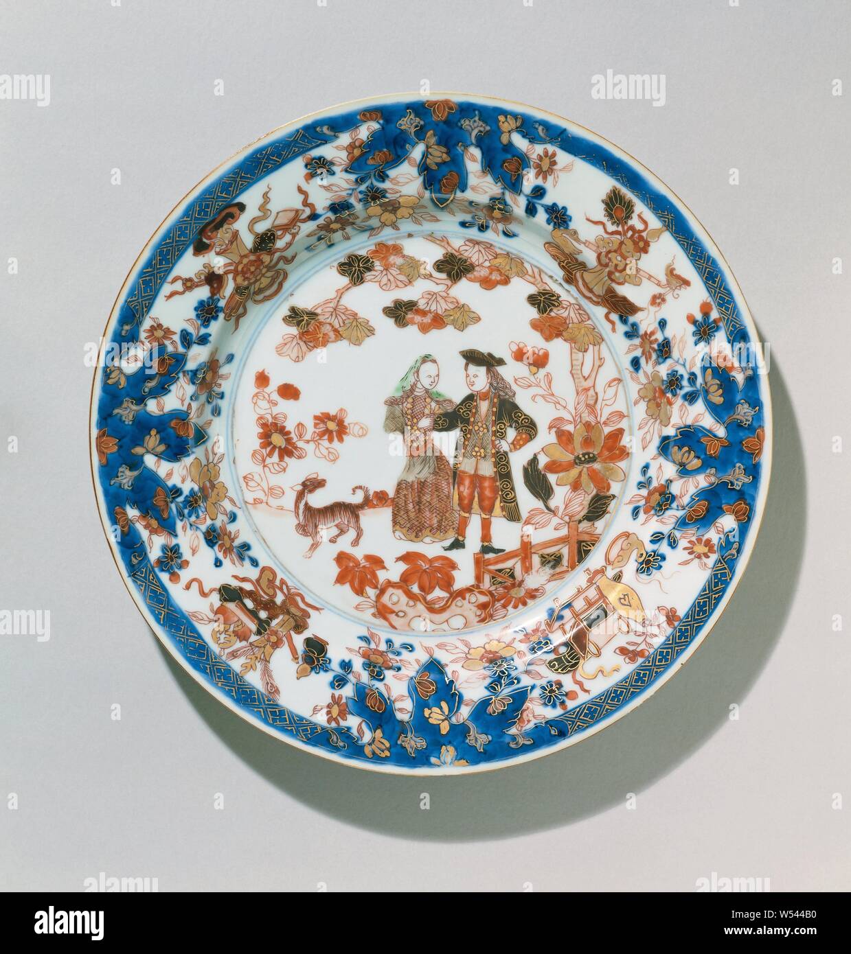 Plate with a Dutch couple and a dog in a fenced garden, Porcelain plate, painted in underglaze blue and on the glaze red, green, black and gold. On the front a representation of a walking Dutch couple with a dog in a fenced garden with a tree, rock and flowering plants, on the wall and border four groups of lucky symbols (including a vase with flowers, coral or peacock feathers, ruyi scepter, fruit basket, scroll, diamond, artemisia leaf), the border with a band with napkin work interspersed with lotus flowers in leaf-shaped boxes. The back with two flower sprays. European representation in Stock Photo