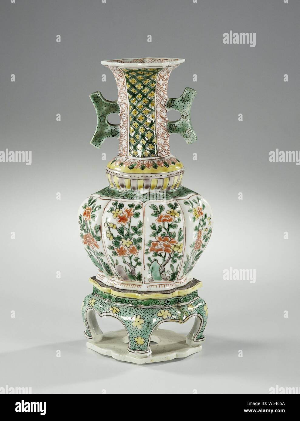 Octagonal vase with two handles, flowering plants and diaper pattern on a separate stand, Octagonal vase of porcelain with two ears standing on the neck, painted on the glaze in red, green, yellow, eggplant and black. The belly is divided into eight compartments with flowering plants near a rock. Above the belly an annular thickening with two bands with leaf motifs. The neck with vertical bands with napkin work. On the ears loose flowers and black dots. The inner edge with a band hatching. The base of the pedestal with yellow flowers and black dots, above it a band with curl. The top Stock Photo