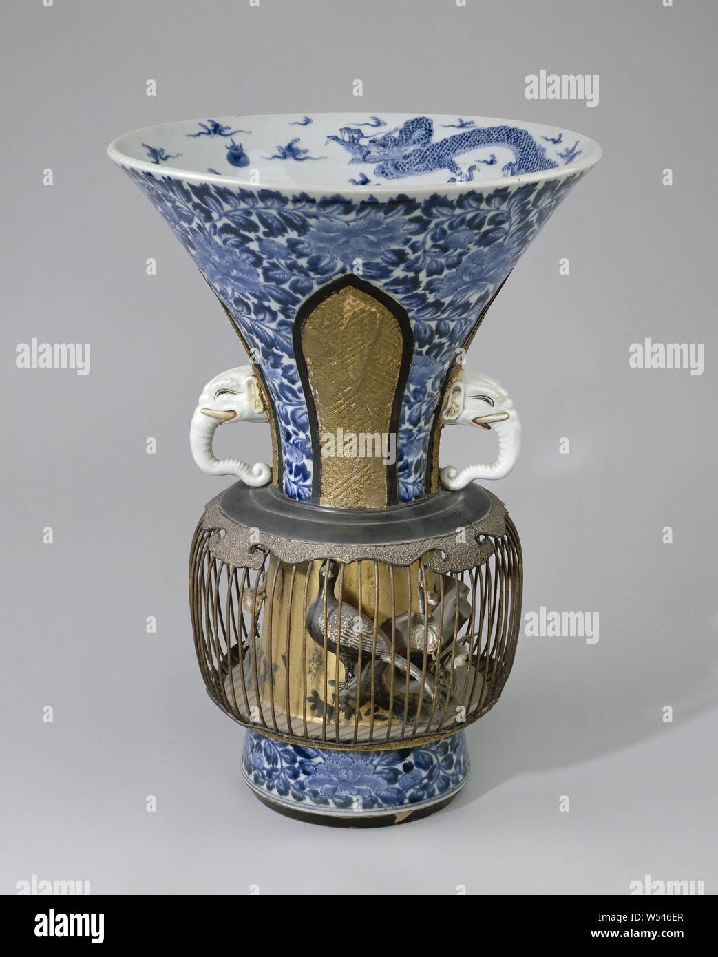 Trumpet-shaped beakervase with a metal birdcage, Trumpet-shaped porcelain vase, painted in underglaze blue and on the enamel red, black and gold. On the wall a pattern of peonies interrupted by four recessed, leaf-shaped boxes with a swastika or grainy pattern in gilt paper mâché. An ear in the shape of an elephant's head in two compartments. A metal bird cage has been placed under these ears around the vase, resting on varnished wooden edges. Below the top edge a band with four ruyi motifs of gilt paper mâché. Rocks, branches and a pair of male and female green pheasant (Phasianus versicolor Stock Photo