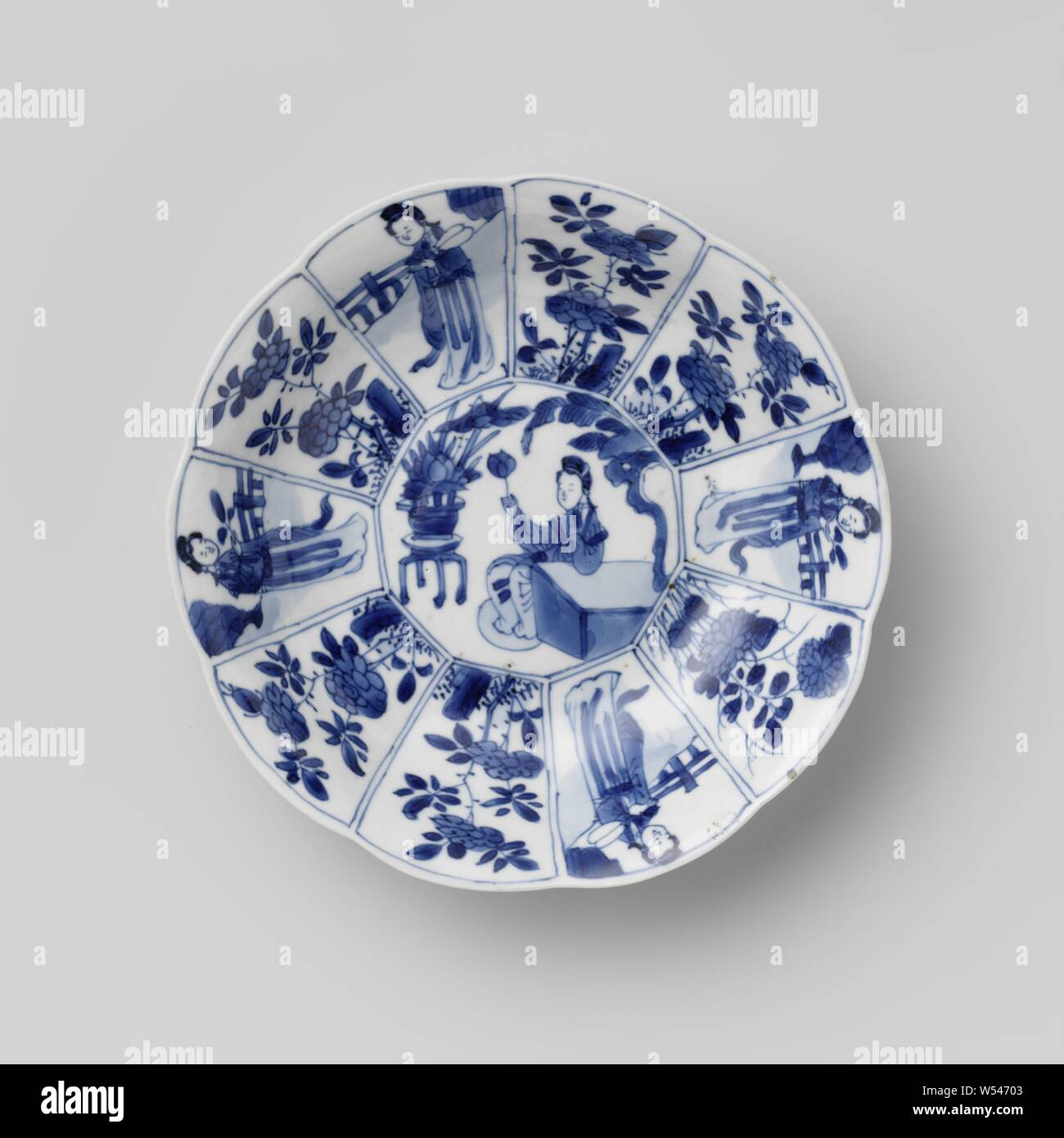 Saucer dish with Chinese ladies in a fenced garden and flowering plants, Porcelain dish with lobed edge, painted in underglaze blue. The wall is modeled in ten compartments. On the shelf a ten-sided medallion with a Chinese lady (long list) sitting at a table with a lotus flower in her hand, behind her a lotus in a pot on a stool. The wall is divided into ten radiating compartments with alternately a lady with a fan or a branch in an enclosed garden or flowering plants. The outer wall with a continuous flower branch. Marked on the bottom with 'yù', jade, in a double circle. Blue and white Stock Photo