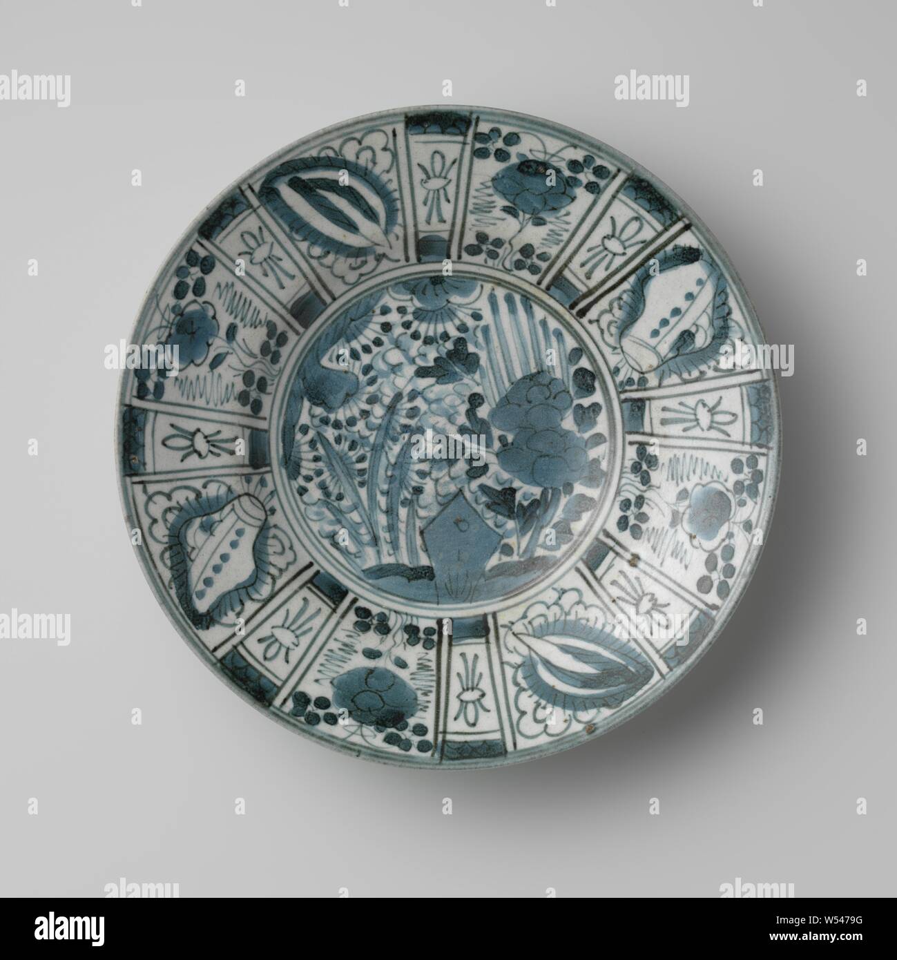 Plate Dish with flowering plants near a rock and a panel decoration, Deep porcelain plate with flat edge, painted in underglaze blue with on the shelf a dense decoration of plants with a stylized rock, on the border surfaces with brush motifs and alternating flowers and valuables. Four proen on the bottom, chip in the foot ring. Decoration following the Chinese kraak porcelain. Blue White., anonymous, Japan, c. 1700 - c. 1799, Edo-period (1600-1868), porcelain (material), glaze, cobalt (mineral), vitrification, h 5.5 cm d 27.3 cm d 12.4 cm Stock Photo