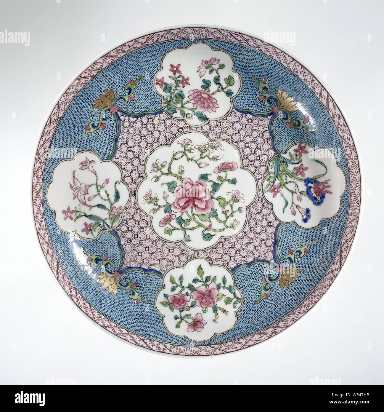 Saucer-dish with flower sprays in shaped panels on a diaper-pattern ground, Porcelain dish covered with a pink glaze and painted on the glaze in blue, red, pink, green, black and gold. On the front of the dish a lobed cartouche with a peony plant in a square scalloped cartouche with napkin, on the corners a stylized lotusrank, on each side of the square a smaller lobed cartouche with a flowering plant (peony, chrysanthemum, iris, balloon bell) against a ground of napkin work, a band with napkin work on the inner edge, the outside covered with a pink glaze. Famille rose with a ruby back Stock Photo
