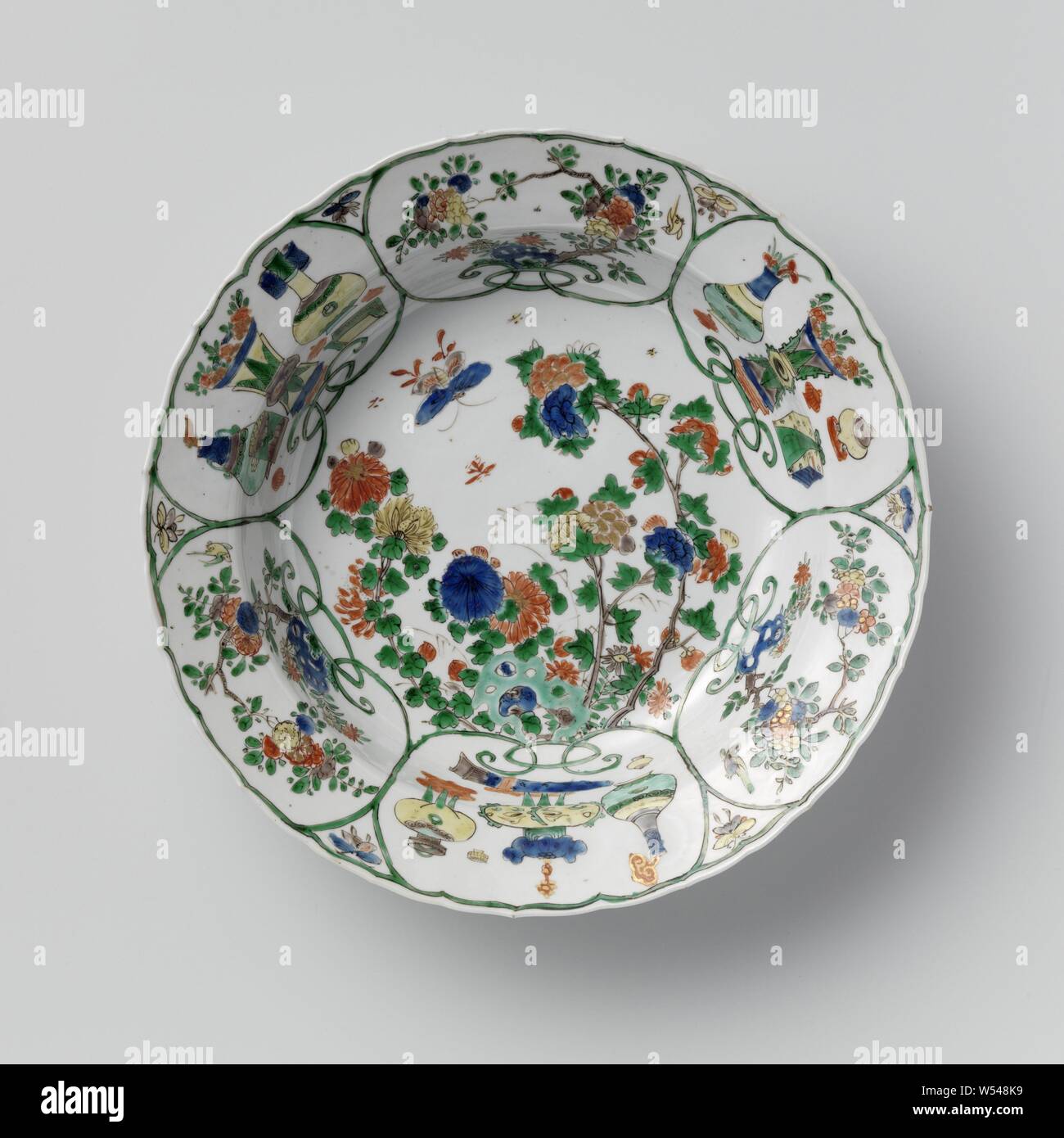 Deep dish with flowering plants near a rock and antiquities, Deep dish of porcelain, painted in blue, red, green, yellow, eggplant, black and gold. On the flat flowering plants (peony, chrysanthemum) and a butterfly near a rock. Six cartouches on the wall and border with alternating flowering plants near a rock and antiques (flower vase, tripod, incense burner, ruyi scepter, books). The outer edge with four flower tendrils, the outer wall is divided into four compartments with a ruyi motif. Marked on the bottom with 'Yù' in a double circle. Famille verte., anonymous, China, c. 1700 - c. 1724 Stock Photo
