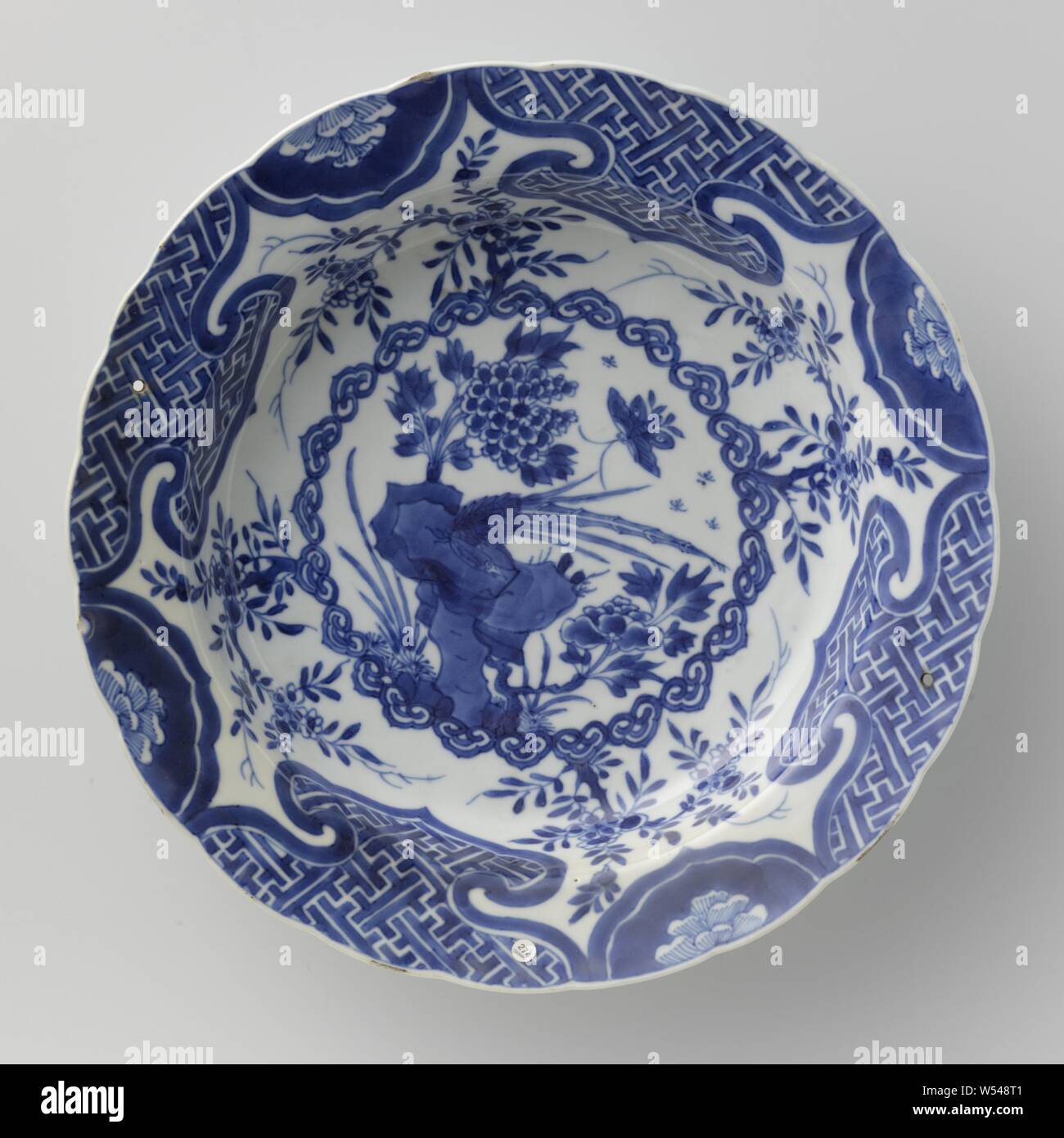 Dish with a pheasant on a rock, butterfly and flower sprays, Deep porcelain dish with lobed edge, painted in underglaze blue. On the shelf a medallion of ruyi motifs containing a pheasant on a rock with two flowering plants, butterfly and insects, a flower branch four times around the medallion, the border with alternating a lobed cartouche with a flower in reserve and a larger cartouche with a geometric pattern, the back with three flower sprays. Two drill holes and a chip in the edge. Blue and white., Battle, fighting in general (naval force), warfare, military affairs (sailing ships Stock Photo