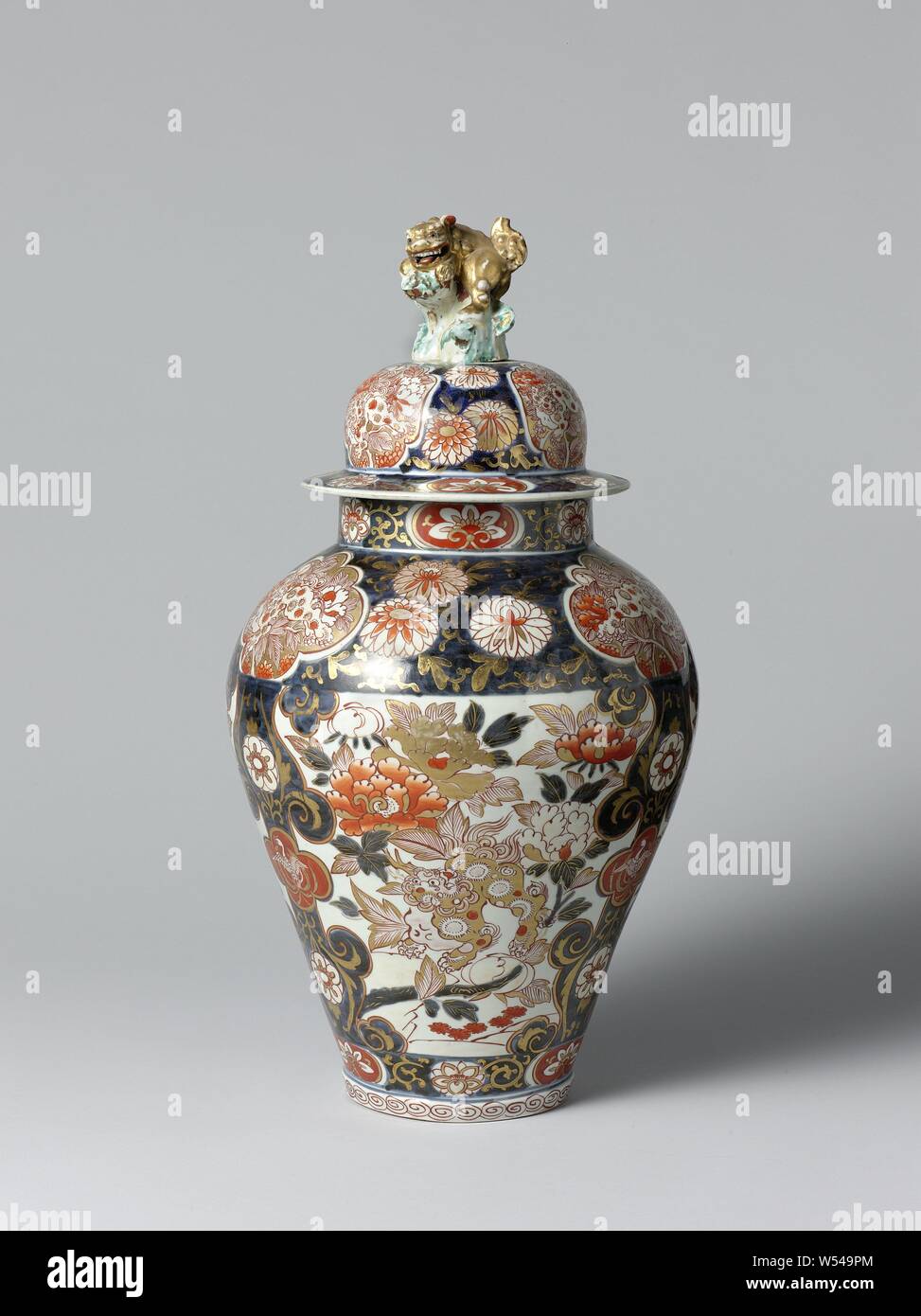 Covered jar with shishi, hoo-birds, flowering plants and floral scrolls, Baluster-shaped porcelain pot with a lid, painted in underglaze blue and on the glaze red, black and gold. On the belly three large cartouches with a shishi between a flowering peony plant, the cartouches are separated by bands of curl work containing a lobed cartouche with a hay bird surrounded by flower vines. On the shoulder a broad band with chrysanthemum drinks interrupted by lobed cartouches with a shishi (lion dog) between a plant. A band with lotus vines around the foot and neck. A band with curl on the foot. Stock Photo