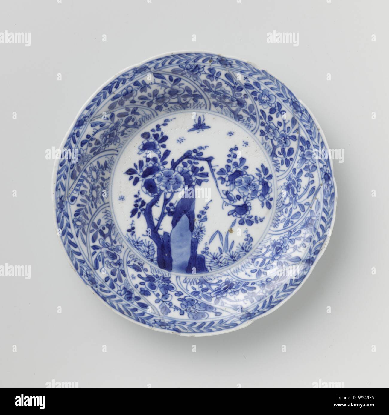 Saucer with twisted panels and flower sprays, Porcelain dish with round wall modeled in ten twisted boxes, painted in underglaze blue. On the flat a rock with a flower branch, plants and butterfly, a flower branch on the wall in each compartment, the border with a flower vine, on the back also a flower branch in each compartment. Marked on the bottom with the Chinese character 'Fú' in a double circle. A few chips in the edge. Blue White., Fú’, anonymous, China, c. 1680 - c. 1720, Qing-dynasty (1644-1912) / Kangxi-period (1662-1722), porcelain (material), glaze, cobalt (mineral), vitrification Stock Photo