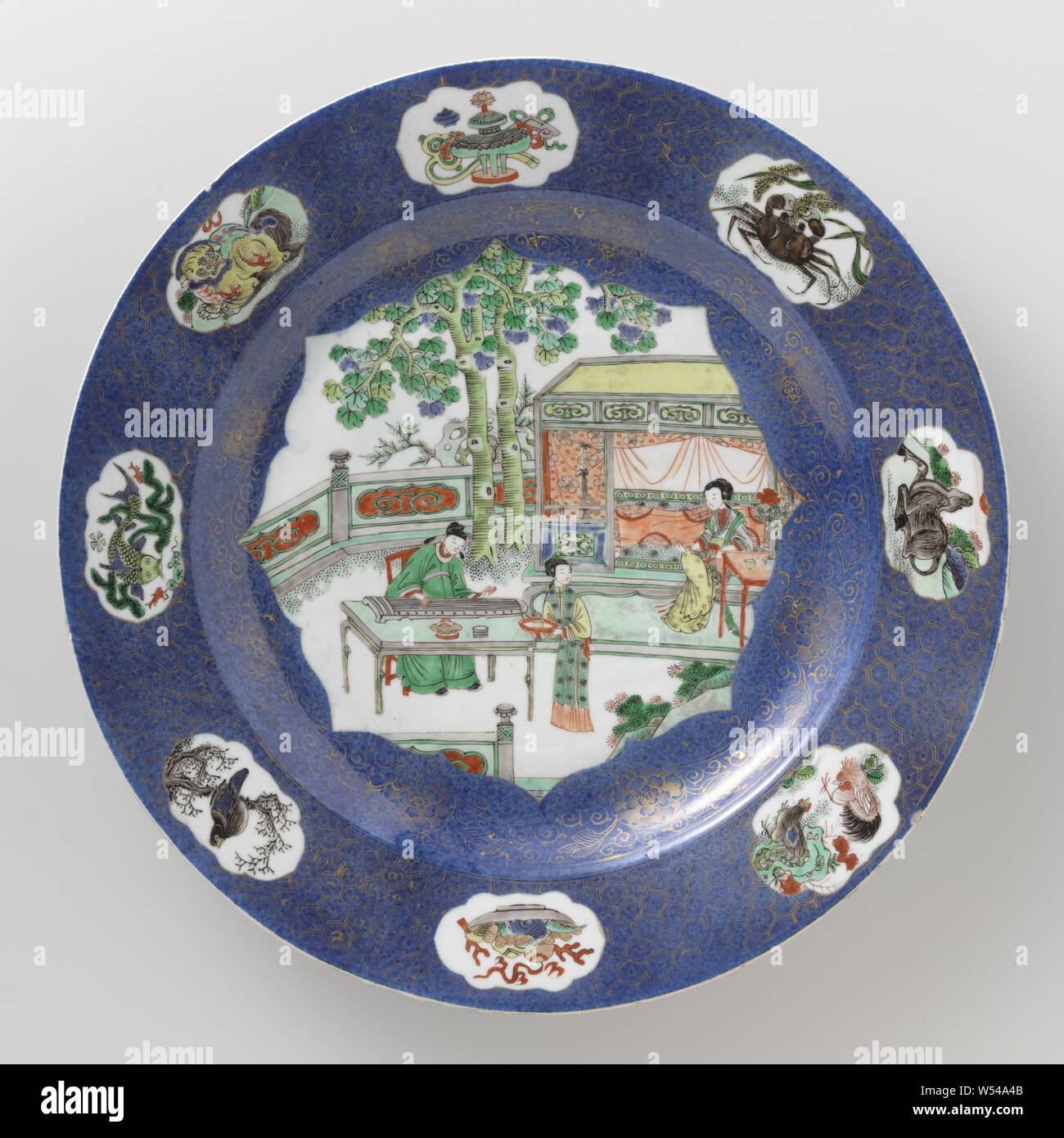 Dish, Dish with bleu poudre and figures on a terrace, Porcelain dish with raised wall and bevel, decorated in underglaze blue and on the glaze blue, red, green, yellow, eggplant, black and gold. The front of the dish is covered with bleu poudré (powder blue) with a large, sculpted field filled with three people on a flat terrace with rocks and two trees, a scholar at a table playing on a qin, a lady sitting at a table in front of a bed, a maid with a bowl on a tray, the wall with a gold decoration on the blue with flower rosettes and tendrils, the border with a gold decoration on the blue Stock Photo