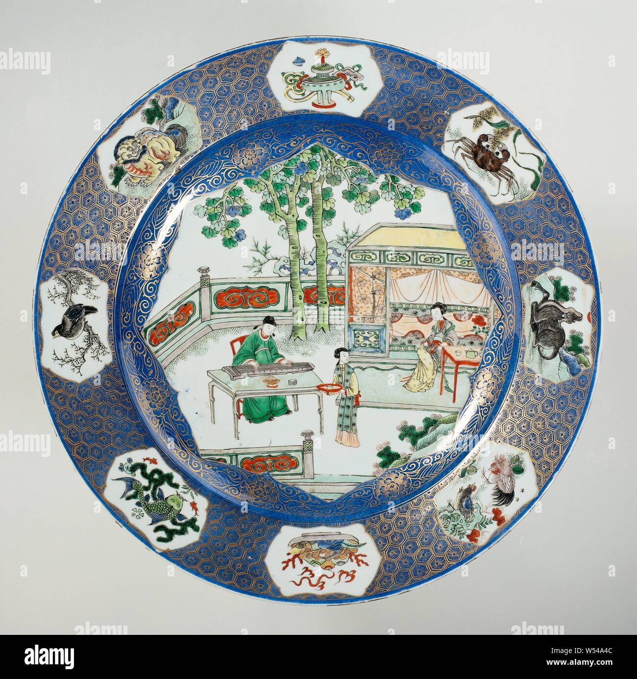 Dish with bleu poudré and figures on a terrace, Porcelain dish with raised wall and bevel, decorated in underglaze blue and on the glaze blue, red, green, yellow, eggplant, black and gold. The front of the dish is covered with bleu poudré (powder blue) with a large, sculpted field filled with three people on a flat terrace with rocks and two trees, a scholar at a table playing on a qin, a lady sitting at a table in front of a bed, a maid with a bowl on a tray, the wall with a gold decoration on the blue with flower rosettes and tendrils, the border with a gold decoration on the blue Stock Photo