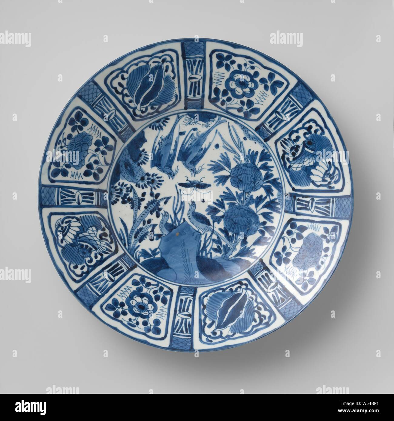 Dish with birds and a butterfly near rocks and flowering plants, Porcelain dish with a wide rim, painted in underglaze blue. On the shelf two flying birds and a butterfly, two more birds on a rock surrounded by flowering plants. The wall and edge is divided into alternately wide and narrow compartments with flower sprays and lucky symbols. The outer wall with three flower sprays. Marked on the bottom with a seal mark. Decoration following the Chinese kraak porcelain. Three proen on the undersides. Blue White., anonymous, Japan, c. 1650 - c. 1700, Edo-period (1600-1868), porcelain (material Stock Photo