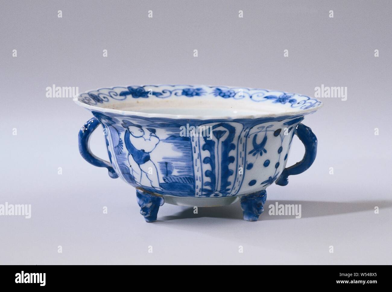 Covered bowl or butter tub with a figure in a landscape, lotus scrolls and flower sprays, Lid bowl or butter dish of porcelain with two ears and on three legs in the form of a monster's head, painted in underglaze blue. The outside of the bowl is divided into wide and narrow compartments, alternately in the wide compartments a stylized person in a landscape and lotus vines, the narrow boxes with a flower branch. A peony branch on the bottom. The lobed border with flower vines. The decoration of the bowl is based on the Chinese kraak porcelain. Two legs have been broken off. Blue White Stock Photo